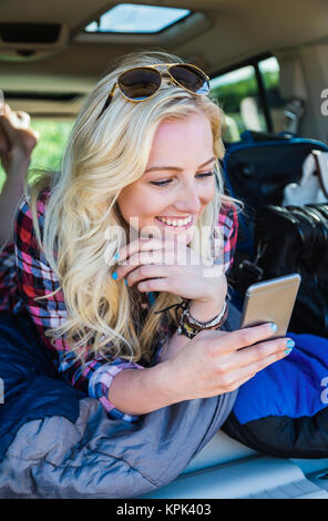 Une jeune femme sur un voyage par la route s'étend dans l'arrière d'un véhicule avec un sac de couchage à l'aide de son téléphone intelligent ; Edmonton, Alberta, Canada Banque D'Images