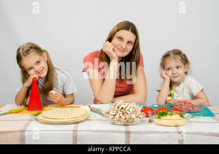 Mère satisfait avec deux filles assis reposant sa tête dans ses mains à la table avec les produits pour pizza Banque D'Images