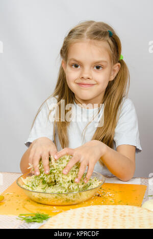 La jeune fille à la table de la cuisine avec un léger sourire se mélange les mains dans un bol le fromage râpé avec des herbes Banque D'Images