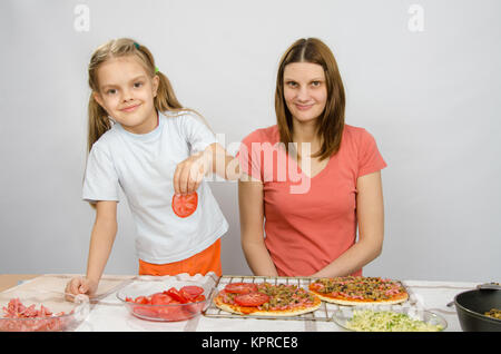 Fille de six ans met sur la pizza tomates sous la supervision de maman Banque D'Images