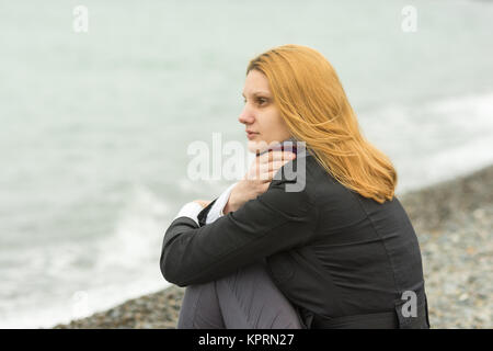 Une fille est assise sur une plage de galets de la mer sur un jour nuageux par temps froid, couvrant le cou foulard