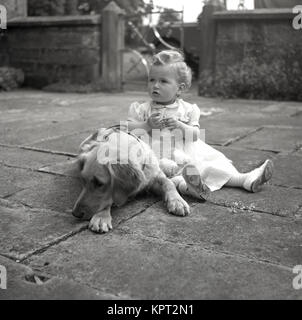 Années 1950, historique, petite fille assise à l'extérieur sur une terrasse ou petite cour avec le chien de la famille. Banque D'Images