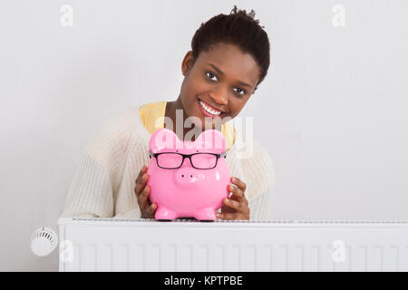 Young African Woman Leaning On radiateur avec Pink Piggybank à la maison Banque D'Images