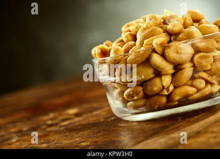 Cuvette ronde en verre transparent rempli de salé huileux pelées arachides rôties sur une table en bois brun, oblique close up Banque D'Images