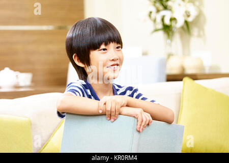 7 ans, petit garçon asiatique sitting on sofa holding a book en souriant. Banque D'Images