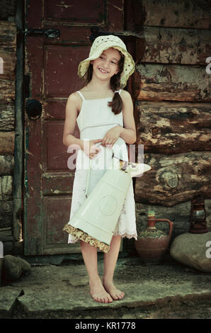 Jeune fille 5-7 ans tenant une cruche et debout devant une vieille maison en bois. Retro et vintage à image. Banque D'Images