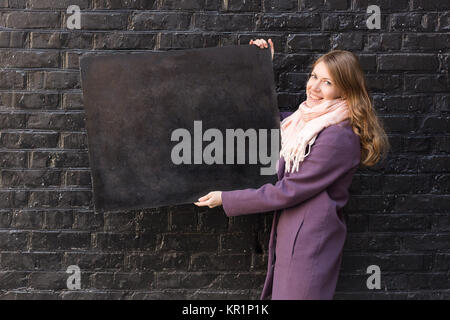 Les arts et l'artisanat, la créativité, l'advertisment concept. adorable femme portant des vêtements d'automne, elle sourit et holding tableau noir sur l'arrière-plan du mur de briques peint Banque D'Images