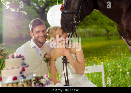 Jeunes mariés restent debout avec un magnifique cheval noir sur une prairie d'été et les raies de l'appareil photo, l'épouse de porter sa robe blanche et tenant un bouquet de mariée, l'époux portant un gilet beige. Banque D'Images