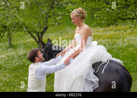 Mariage à la campagne. La mariée dans sa robe de mariée blanche est assis sur un cheval foncé et son époux lui permet de démonter quand le cheval Banque D'Images