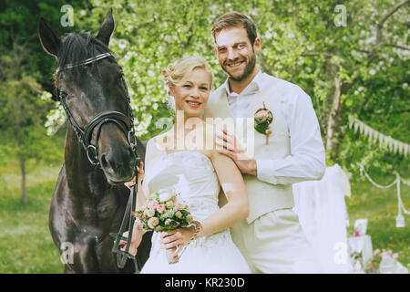 Jeunes mariés restent debout avec un magnifique cheval noir sur une prairie d'été et les raies de l'appareil photo, l'épouse de porter sa robe blanche et tenant un bouquet de mariée, l'époux portant un gilet beige. Banque D'Images