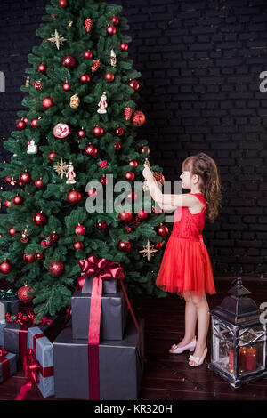 Jolie petite fille en robe courte rouge se tient près de l'immense arbre de Noël décoré avec des ornements d'or et rouge pour des fêtes. De nombreux présente u Banque D'Images