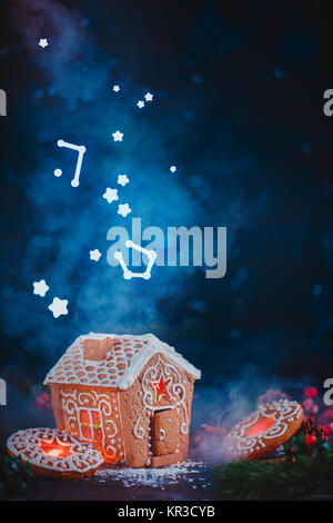 Gingerbread House avec windows brillant, l'augmentation de la fumée d'une cheminée et de constellations dans une scène de nuit avec le ciel étoilé. Noël Nourriture photographie Banque D'Images