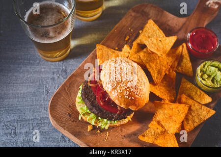 Portrait sur burger fraîchement préparé dans sesame bun, croustilles, la salsa et le guacamole sur une planche à découper avec de la bière sur la table Banque D'Images