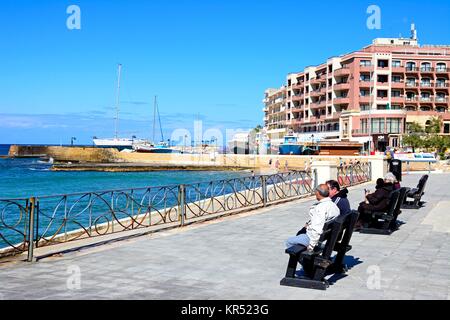 Les touristes assis sur des bancs le long de la promenade avec vue sur plage et Port, Marsalforn, Gozo, Malte, l'Europe. Banque D'Images