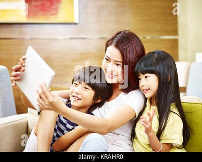 Young Asian mother and her son and daughter sitting on couch prenant un using digital tablet selfies. Banque D'Images