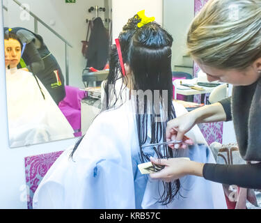 Femme avec de longs cheveux noirs lui coupe les cheveux mouillés en arrière recouvert d'une cape salon blanc et assis en face d'un miroir. Le coiffeur utilise des ciseaux. Banque D'Images