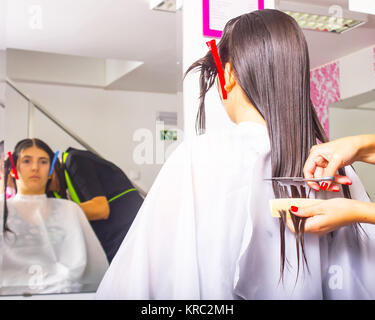 Femme avec de longs cheveux noirs lui coupe les cheveux mouillés en arrière recouvert d'une cape salon blanc et assis en face d'un miroir. Le coiffeur utilise des ciseaux. Banque D'Images