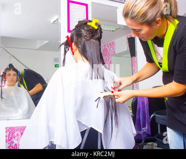 Femme avec de longs cheveux noirs lui coupe les cheveux mouillés en arrière recouvert d'une cape salon blanc et assis en face d'un miroir. Le coiffeur utilise des ciseaux. Banque D'Images