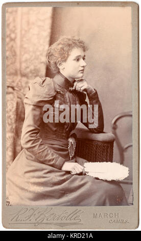 Une jeune femme pose victorienne dans le studio du photographe, vêtue d'une robe élégante avec un haut col et broderie sur le corsage et des manches. Elle est assise de profil, avec une plume blanche ventilateur dans sa main. Date : vers 1890