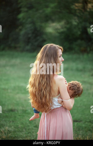 Jeune belle mother hugging son petit petit garçon contre l'herbe verte. Femme heureuse avec son petit garçon sur une journée ensoleillée d'été. Balades en famille sur le pré. Banque D'Images