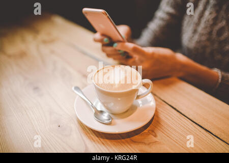 Mains près d'une belle jeune fille utilise,texte sur un téléphone mobile à une table en bois près d'une fenêtre et boit du café dans un café décoré avec des décorations de Noël. Vêtu d'un chandail tricoté gris Banque D'Images