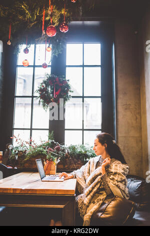 Belle jeune fille utilise la technologie de l'ordinateur portable, les types de texte le moniteur dans un café au bord de la fenêtre, à la table en bois en hiver, décoré avec des décorations de Noël.vêtu d'un pull en laine tricoté gris Banque D'Images