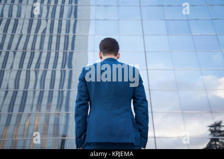Vue arrière de l'homme d'affaires en costume chic debout avec les mains dans les poches sur fond de verre. Banque D'Images