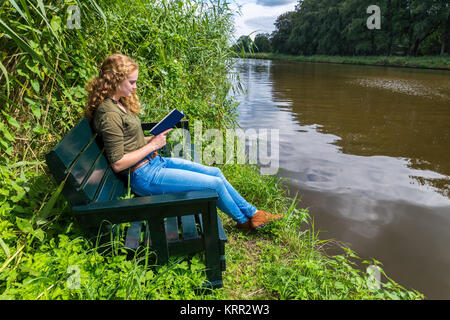 Young caucasian woman reading book sur banc en bois à la rivière Banque D'Images