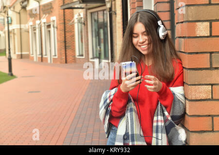 Smiling Asian Girl standing près du mur et écouter de la musique sur un casque, de tenir le téléphone dans la main et regarder Banque D'Images