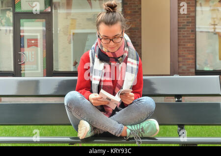 Fille asiatique dans les verres est assis sur un banc, croise ses jambes et lit un livre. Autour de la rue et le bâtiment Banque D'Images