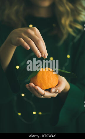 La mandarine brut frais d'agrumes avec des feuilles dans les mains du blond girl wearing robe vert avec lumières de bokeh, selective focus, copiez l'espace. Winte Banque D'Images