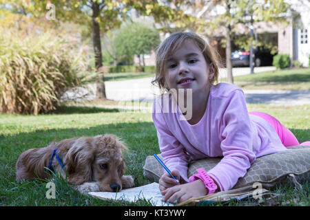 Jeune fille et son chiot cocker de détente en plein air, sur l'herbe te avec un coloriage de livre comme elle sourit joyeusement à l'appareil photo Banque D'Images