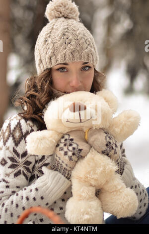 Beau, agréable, souriant et portant peu,sweater hut et mitaines et petite fille avec un ours dans la forêt d'hiver.Portrait de jolie fille avec mignon,joli jouet.Cute,belle,belle fille hug a fluffy,blanc beige ours.Girl et l'ours en hiver Banque D'Images