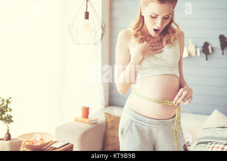 Grande taille. Surprise agréable pregnant woman holding type de mesure et de l'utiliser sur son ventre tout en exprimant l'étonnant Banque D'Images