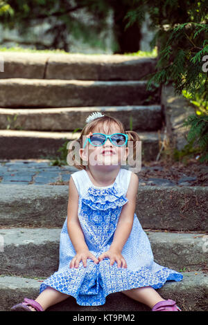 Petite fille portant des lunettes de soleil est assis sur les étapes et les sourires à la recherche le ciel Banque D'Images