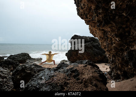 Femme en manteau de pluie faisant reposer sur l'asana yoga plage sur l'océan pendant une tempête. Concept de voyage yoga sur Bali Banque D'Images