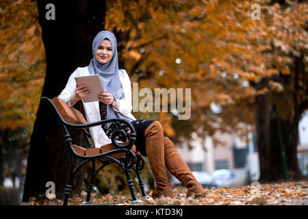 Belle jeune fille de porter le hijab en couleurs de l'automne. À l'aide de tablette sur un banc de parc Banque D'Images