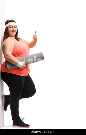 Portrait d'une femme en surpoids avec un tapis d'exercice faisant un signe du pouce vers le haut et appuyé contre un mur isolé sur fond blanc Banque D'Images