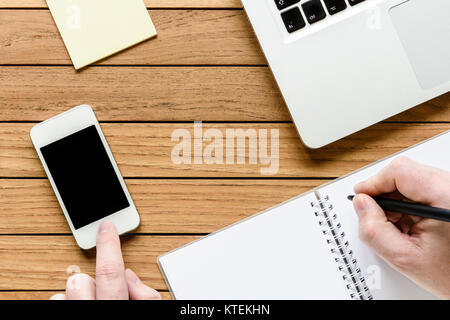 Vue de dessus de bureau en bois, un homme avec la main sur un ordinateur portable et de toucher un smartphone, un ordinateur portable et le bloc-notes. Banque D'Images