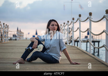 Porter Féminin décontracté skinny jeans bleu foncé et chemise bleue, assis au quai avec plancher en bois et tenant un bouqet de oeillets blancs Banque D'Images