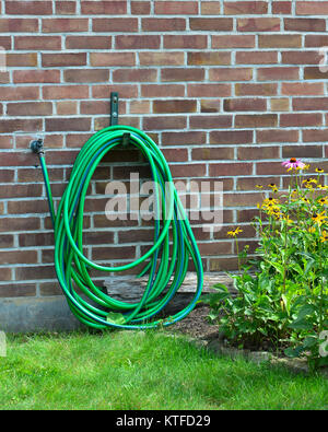 Tuyau de jardin vert et bleu enroulé sur un cintre à côté d'une pelouse et un jardin de fleurs contre un mur de brique rouge. Banque D'Images