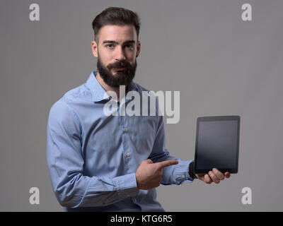 Jeune homme barbu sceptique doigt montrant l'écran de l'ordinateur tablette blanc sur fond gris studio. Banque D'Images