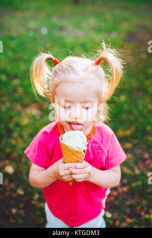 Peu drôle Caucasian girl blonde aux yeux bleus avec deux queues sur la tête de manger une glace dans un cornet gaufré bleu tasse de assis sur l'herbe verte.tous confrontés à d Banque D'Images