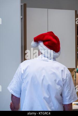 Homme plus âgé en chef les blancs et Santa hat à la recherche d'un frigo dans une cuisine à l'époque de Noël Banque D'Images