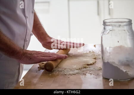 Close up de plus dans les blancs chef pâte de roulement avec un rouleau à pâtisserie faire sur un bloc de bois avec un pot de farine dans une cuisine Banque D'Images