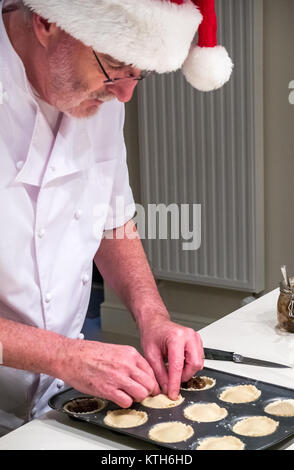 Homme plus âgé en chef les blancs et Santa hat remplissant mince pie cas avec la viande hachée dans un moule prêt pour la cuisson à l'époque de Noël dans une cuisine Banque D'Images