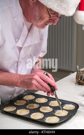 Homme plus âgé en chef les blancs et Santa hat remplissant mince pie cas avec la viande hachée dans un moule prêt pour la cuisson à l'époque de Noël dans une cuisine Banque D'Images