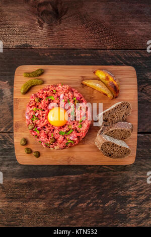 Une photo prise à la verticale d'un steak tartare d'un jaune d'œuf cru, cornichons, câpres, pain de seigle, pommes de terre rôties, tourné à partir de ci-dessus sur une texture rustique foncé avec Banque D'Images