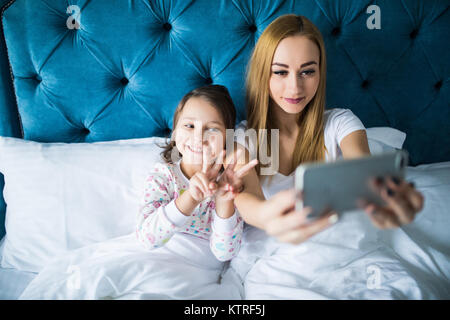 Mother and Daughter lying on bed and taking self portrait avec le smartphone. Femme tenant une petite fille avec des selfies dans la chambre. Banque D'Images