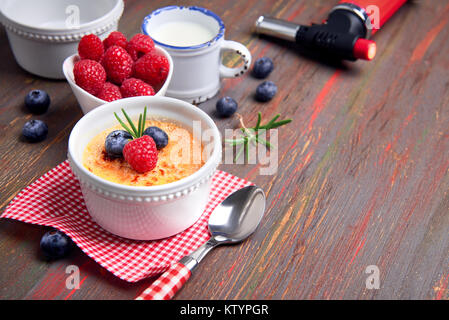 Crème brûlée à la framboise, la myrtille et le romarin en ramequin blanc avec des ingrédients et flambeau ardent sur table en bois rustique Banque D'Images